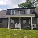 Side view of a remodeled home with gray siding, stone accents, dark roofing, and a balcony.