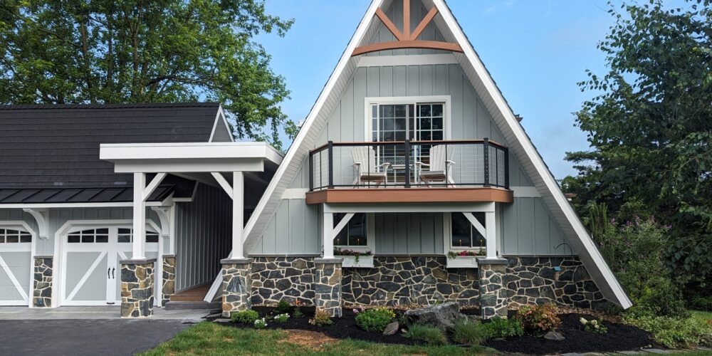 Front exterior of a unique triangle shaped home with new light gray siding, white trim, stone accents, brown balcony, and a matching 2-car garage