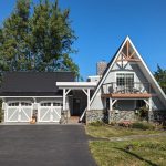 Front exterior of a unique triangle shaped home with new light gray siding, white trim, stone accents, brown balcony, and a matching 2-car garage