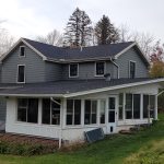 Back and side view of a remodeled home with greenish-gray siding, a white sun room, a brick chimney, new windows, and black roofing.