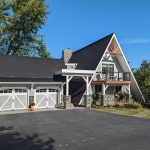Front and left side exterior view of a unique triangle shaped home with new light gray siding, white trim, stone accents, brown balcony, and a matching 2-car garage