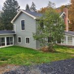 Back and side exterior of a remodeled home with greenish gray siding, new windows, 2 sunrooms, a brick chimney, and a medium sized tree.