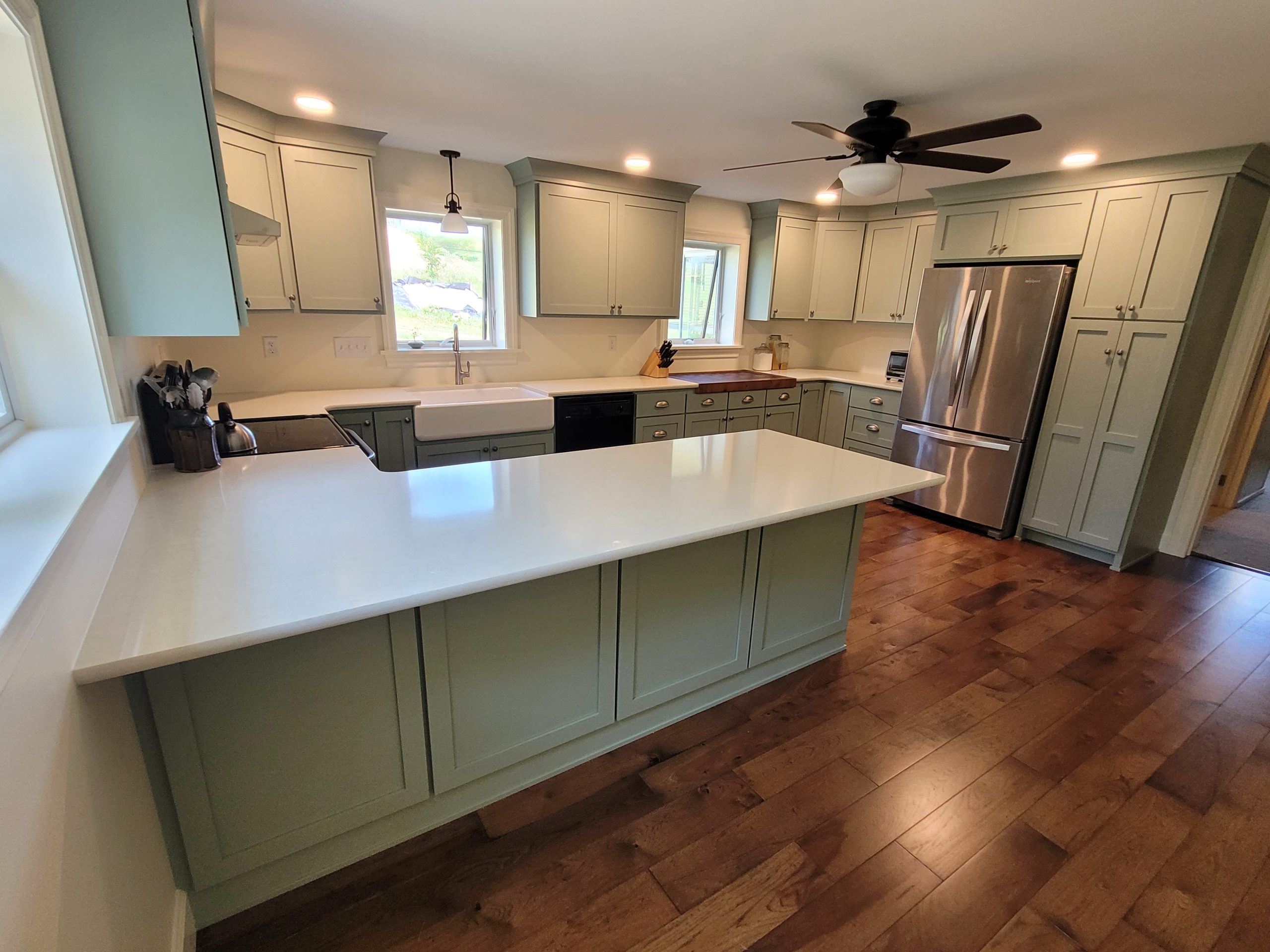 Overview of a renovated kitchen with sage green cabinets, white counter tops, white walls, dark brown hardwood flooring, a stainless steel fridge, a black ceiling fan, and a white farmhouse sink.