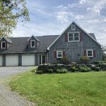 Back exterior view of a home with aged and faded siding, red shutters, white trim, gray roofing, and 3 white garage doors on an attached garage.