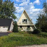 Front side exterior view of a unique triangle shaped home with light yellow wood siding, black roofing, and a light yellow balcony