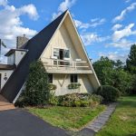 Front and left side exterior view of a unique triangle shaped home with light yellow wood siding, black roofing, and a light yellow balcony