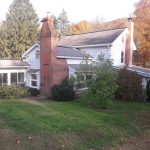 Back and side view of an aged home with 2 brick chimneys, white siding, gray roofing, and large shrubs in the yard.