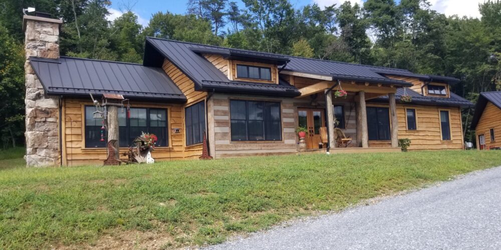 Front exterior view of a custom home with rustic wood siding, black metal roofing, black windows, a front porch with log posts, and a stone chimney.