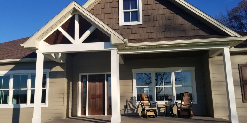 Close up of a new home's covered front porch with an a-frame roof, white posts, tan siding, shake accent siding, white windows, a brown front door, a set of rocking chairs, and a small table.