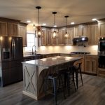 Overview of a new kitchen with brown stained cabinets, speckled granite countertops, a kitchen island with 3 black stools, dark stainless steel appliances. light colored walls, and hardwood flooring.
