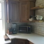 Close up of a remodeled kitchen corner with brown corner cabinets, floating shelves, and kitchen appliances on a white marble countertop.