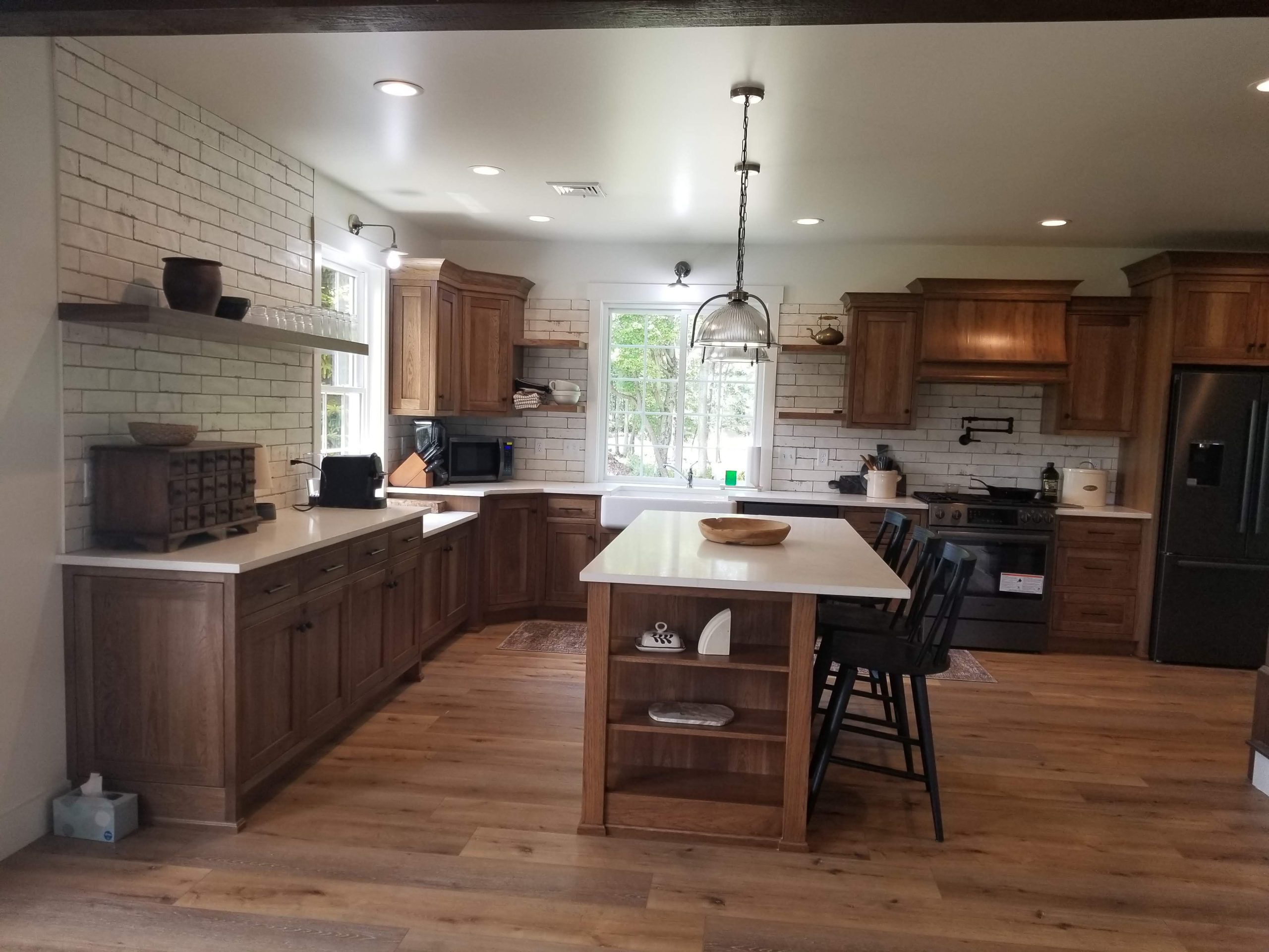 Remodeled kitchen with a brown and white island with black stool chairs, wood flooring, white tile backsplash, brown cabinets, and white marble countertops.