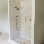 Close up of a new shower in a remodeled bathroom with white tile walls, white tile floors, built in wall shelves, and 2 metallic shower heads.