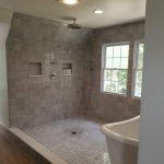 Close up of a shower and tub combination with a white soaking tub, gray tiled walls, and gray tiled shower flooring in a remodeled bathroom with 2 silver shower heads.