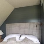 Close up of a bedroom corner with green wood wall cladding, a gray square-patterned headboard, and bed with gray and white bedding.