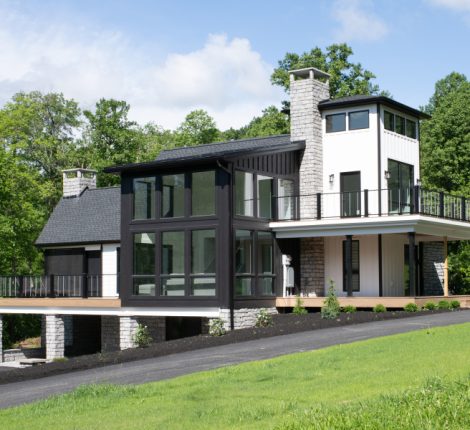 Modern home renovation with a front deck, a covered wrap around porch, a wraparound balcony, 2 stone chimneys, white walls, black windows, and a black accent wall.