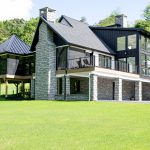 Modern remodel of a home with white and black siding, black trim, black roofing, stone veneer accents, a stone chimney, 2 decks, and a screened-in deck with a black metal roof.