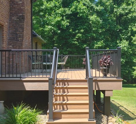New, tan deck with black railings attached to a red brick house in a backyard surrounded by trees.