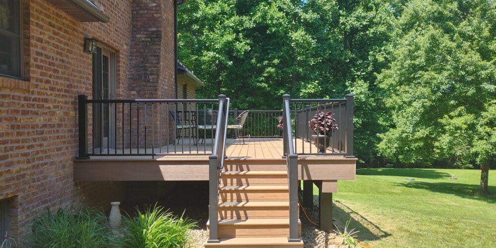 New, tan deck with black railings attached to a red brick house in a backyard surrounded by trees.
