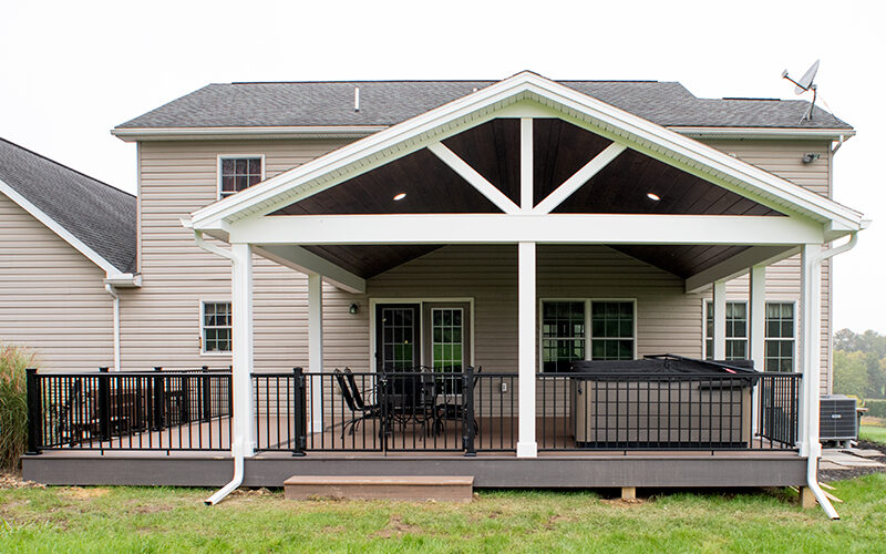 New covered deck with brown decking, white beams and posts, black aluminum railings, a black outdoor dining set, a hot tub, and a brown plank ceiling with puck lights.