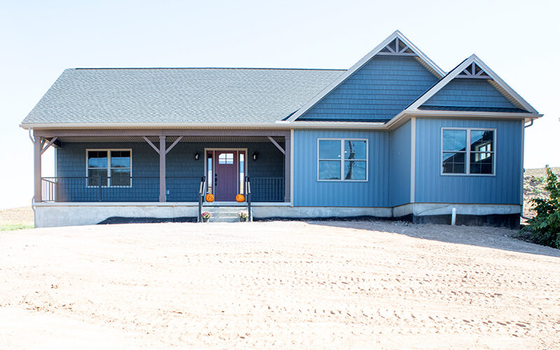 Front exterior of a new ranch-style home with blue siding, decorative blue shakes, white trim, white windows, a gray asphalt roof, and a covered front porch with black aluminum railings.
